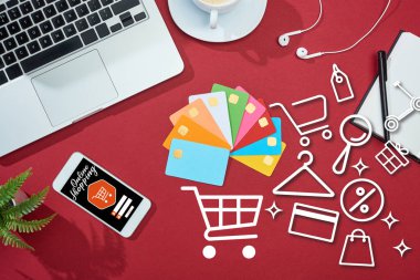 top view of multicolored empty credit cards on red background with online shopping illustration on smartphone near laptop, earphones and coffee