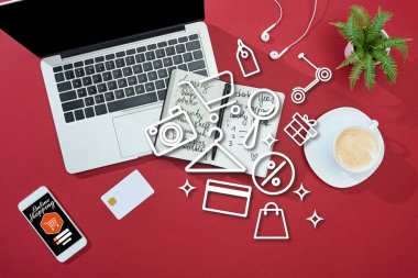top view of credit card on red background with online shopping illustration on smartphone near laptop, earphones, coffee, notebook and plant