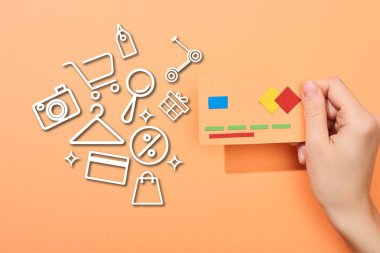 cropped view of woman holding credit card template on peach background with icons illustration