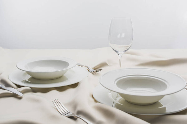 Тарелки со столовыми приборами и прозрачный бокал вина на белой скатерти на сером фоне
