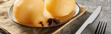 delicious pear in wine with anise and cinnamon served on plate on napkin with knife and fork on grey concrete surface, panoramic shot clipart