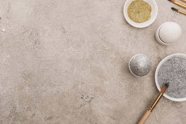 Tavuklu yumurta, gümüş ve altın parıltılı gri beton yüzeyde boya fırçaları.