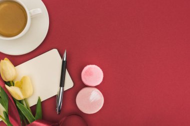 İpek kurdeleli lalelerin üst görüntüsü, kırmızı üzerine izole edilmiş kahvenin yanındaki boş makaronlar ve boş defter.