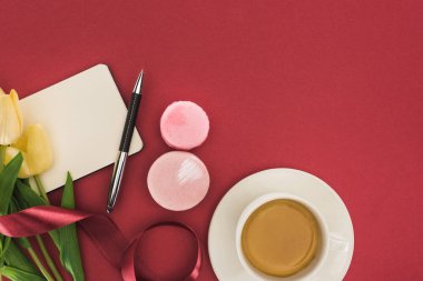 Lalelerin üst görüntüsü ve kahve fincanı, badem kurabiyesi ve kırmızı üzerine izole edilmiş kurdeleli boş bir defter.