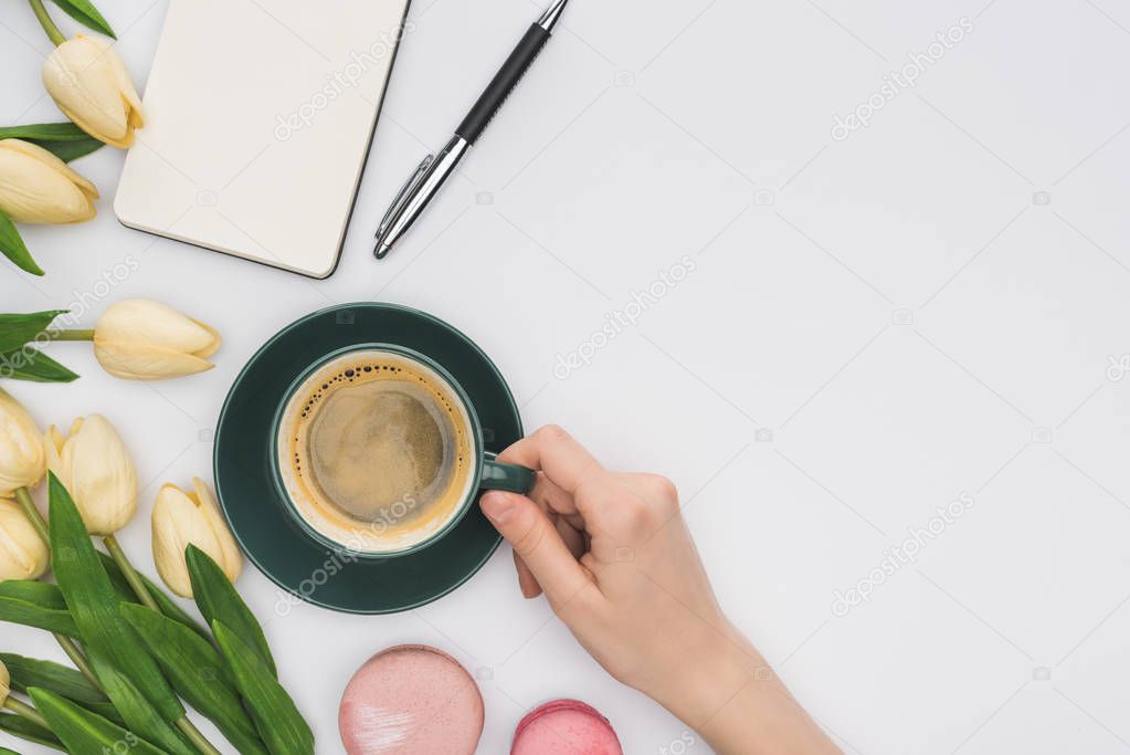 cropped view of woman holding coffee cup near tulips, blank notebook and delicious macarons isolated on white