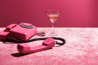 Pembe kadife kumaş üzerinde, klasik telefonun yanında cam içinde gül şarabı. Pembe, kızsı bir konseptte izole edilmiş.