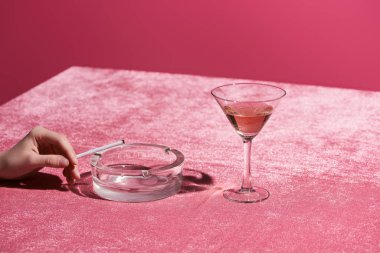 Kül tablasının yanında sigara ve pembe kadife kumaş üzerinde gül şarabı olan pembe, kızsı bir konsepte izole bir kadın manzarası.