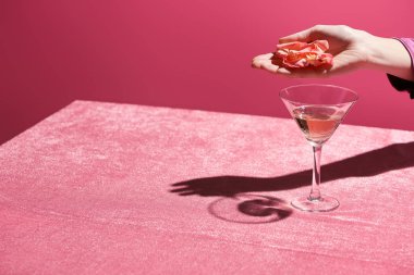 Kırpılmış görüntüde gül yapraklarını cam üzerinde tutarken kadife kumaş üzerinde pembe, kızsı bir konseptte izole edilmiş gül şarabı. 