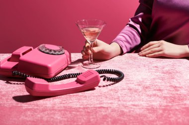Kırpılmış bir görüntü, pembe, kızsı bir konsepte izole edilmiş kadife kumaş üzerinde retro telefonun yanında bir bardak gül şarabı tutan bir kadın. 