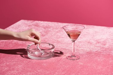 Kırpılmış kadın manzarası pembe, kızsı bir konsepte izole edilmiş kadife kumaş üzerinde gül şarabı bardağının yanında sigara söndürüyor. 