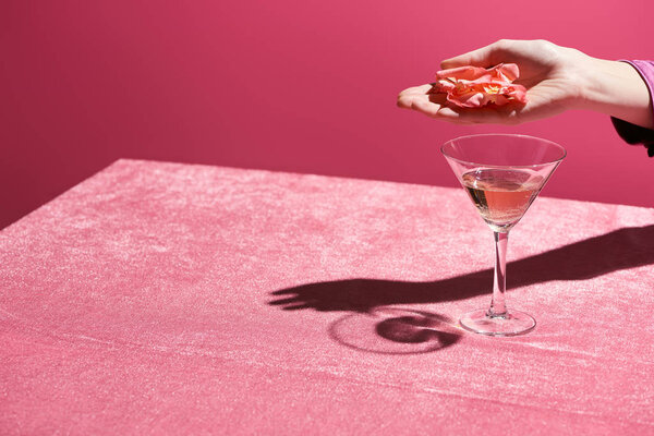 обрезанный вид женщины, держащей лепестки роз над бокалом розового вина на велюровой ткани, изолированной по розовой, девичьей концепции
 