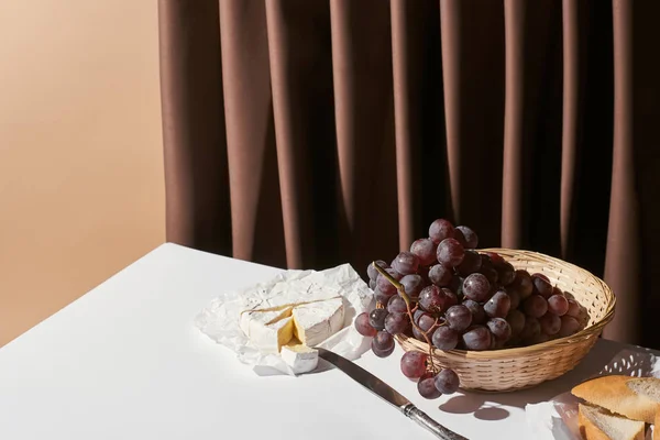 ベージュで隔離されたカーテンの近くのテーブルの上でバスケット ブリーチーズ バゲットのブドウと古典的な静物画 — ストック写真