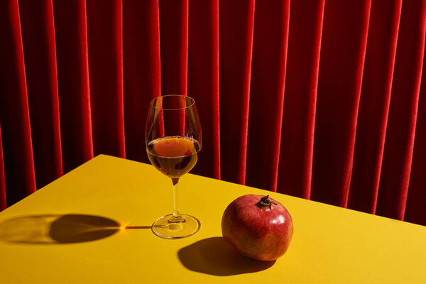 classic still life with pomegranate near glass of red wine on yellow table near red curtain