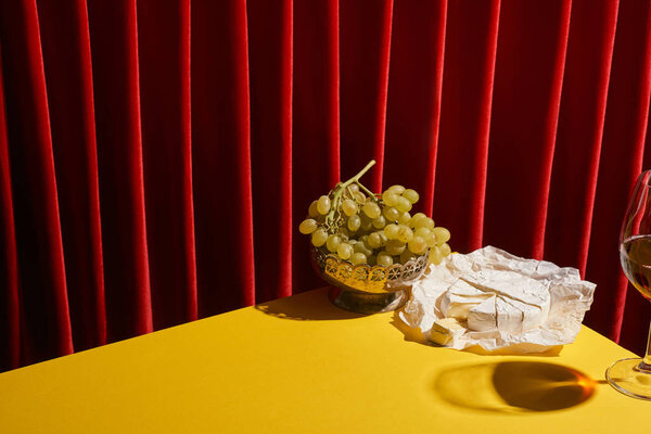 classic still life with Camembert near glass of red wine and grape on yellow table near red curtain