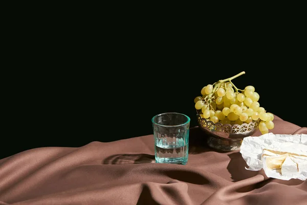 黒で隔離された茶色のテーブルクロスとテーブルの上のガラスでカンベールチーズ ブドウ 水と古典的な静物画 — ストック写真