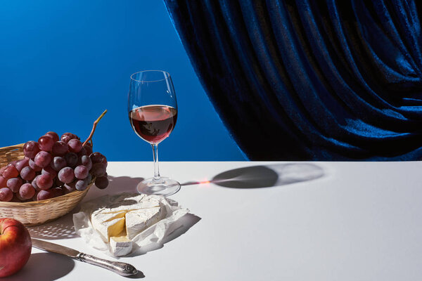 classic still life with fruits, red wine and Camembert cheese on white table near velour curtain isolated on blue