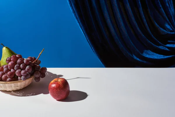 ブルーに隔離されたベロアカーテンの近くの白いテーブルの上のウィッカーバスケットのブドウ リンゴと古典的な静物画 — ストック写真