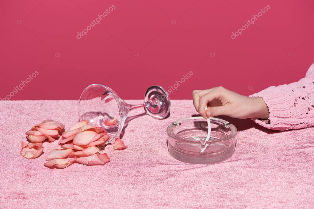 Cropped view of woman putting out cigarette near glass with scattered petals on velour cloth isolated on pink, girlish concept