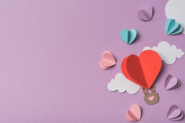 Renkli kağıt kalplerin ve kalp çevresindeki bulutların üst görüntüsü menekşe arkaplanındaki kağıt hava balonu şeklindedir.