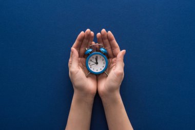 cropped view of woman holding alarm clock on blue background clipart