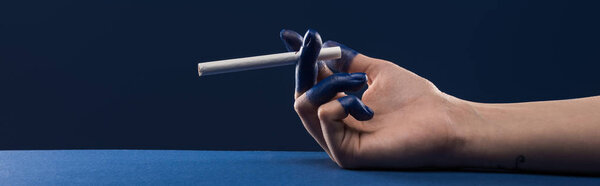 обрезанный вид женской руки с раскрашенными пальцами, держащими сигарету изолированный на голубом, панорамный снимок
