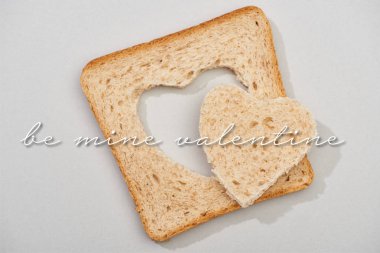Top view of slice of bread with carved heart and be mine valentine lettering on grey background