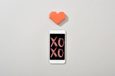 Top view of smartphone with xoxo illustration and paper heart on grey background