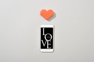 Top view of smartphone with love word and paper heart on grey background