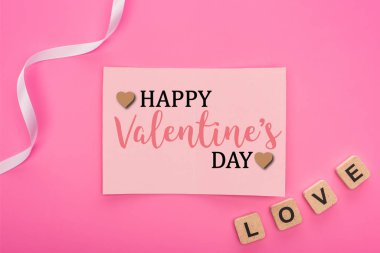 top view of empty paper card with  happy valentines day lettering near ribbon and love lettering on wooden cubes isolated on pink clipart
