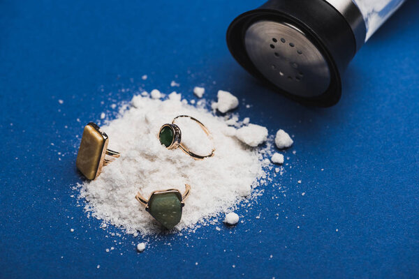Close up view of jewellery rings on salt heap with salt shaker on blue surface