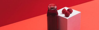 fresh berry juice in glass bottle near ripe raspberries on cube on red background, panoramic shot clipart