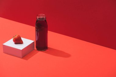 Kırmızı arka planda olgun çileğin yanında cam şişede taze meyve suyu.