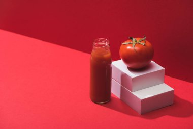 Kırmızı arka planda duran olgun domatesin yanında cam şişede taze sebze suyu.