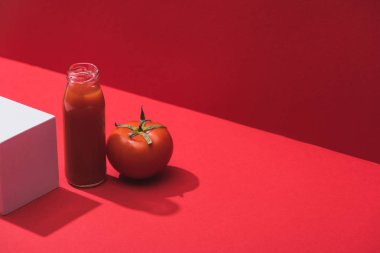 Kırmızı arka planda olgun domates ve küp yanında cam şişede taze sebze suyu.