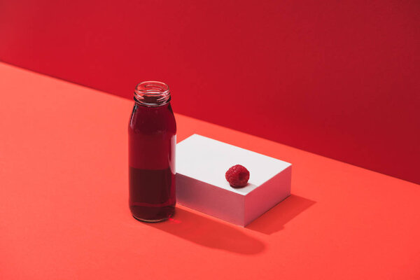 Свежий ягодный сок в стеклянной бутылке возле спелой малины на кубе на красном фоне
