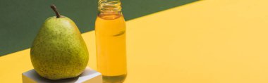 fresh juice in bottle near pear and white cube on green and yellow background, panoramic shot