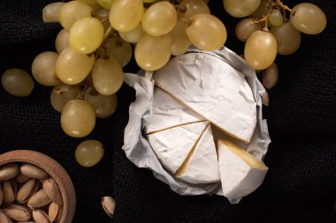 Top view of pieces of camembert with grapes and bowl of pistachios isolated on black  clipart