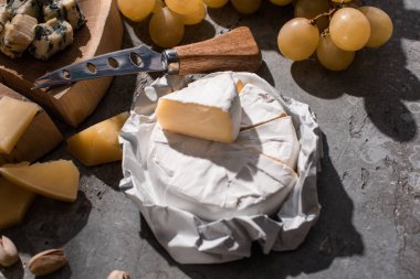 Grana Padano 'nun yanında Camembert peyniri, ahşap tahtada bıçak, üzüm ve gri arka planda fındık.