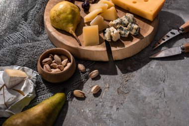 Pieces of cheese with pear and olives on wooden board next to knives, nuts on grey background clipart