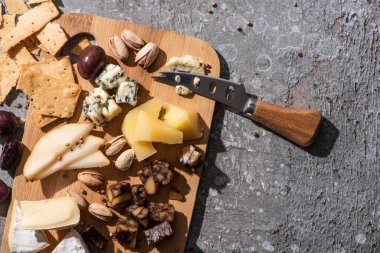 Peynir, zeytin, şam fıstığı, armut dilimleri, kraker ve bıçağın gri arka plandaki kesme tahtasındaki üst görüntüsü.