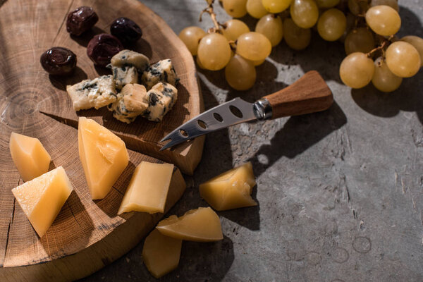 Grana padano and dorblu with olives and knife on wooden board next to grapes on grey background