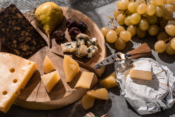 Different kinds of cheese with fruits, olives and knife on wooden board on grey background