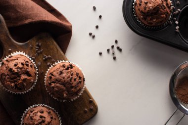 top view of fresh chocolate muffins on wooden cutting board near brown napkin and cocoa powder on marble surface