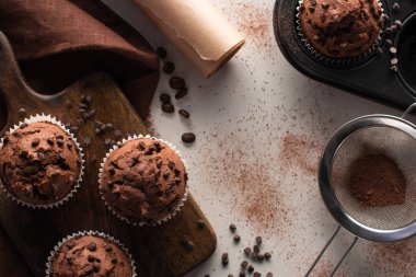 top view of fresh chocolate muffins on wooden cutting board near brown napkin, parchment paper and cocoa powder on marble surface