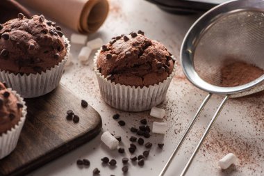 close up view of fresh chocolate muffins near marshmallow, cocoa powder and sieve on wooden cutting board