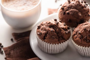 close up view of fresh chocolate muffins on white plate and brown napkin near cappuccino on marble surface