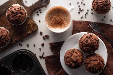 top view of fresh chocolate muffins on cutting board, white plate and brown napkin near fork and cappuccino on marble surface