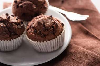 close up view of fresh chocolate muffins on white plate near fork on brown napkin