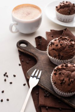 fresh chocolate muffins on wooden cutting board near napkin, fork and coffee