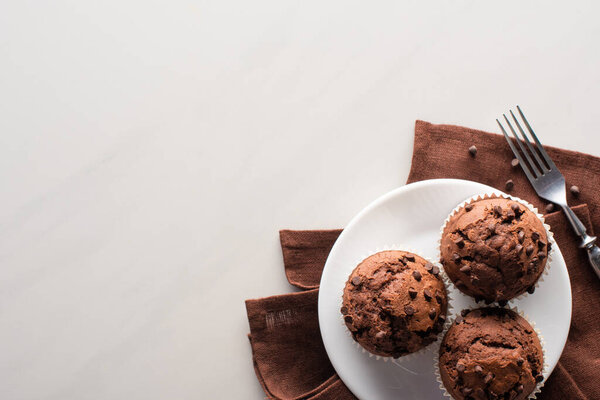 top view of fresh chocolate muffins on white plate and brown napkin near fork on marble surface
